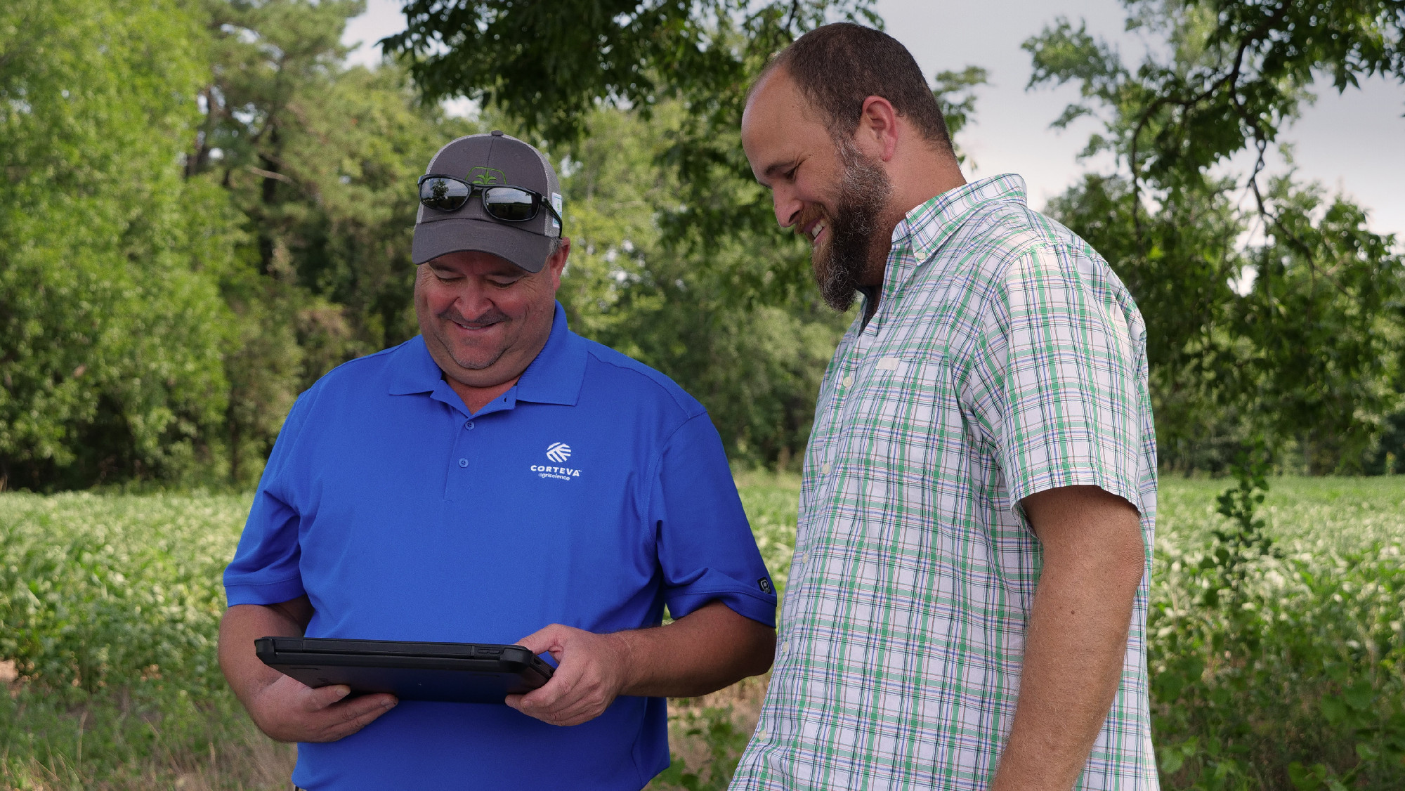 Two men standing outdoors in a field, smiling while looking at a tablet device. One man is wearing a blue polo shirt and sunglasses, and the other is wearing a plaid shirt.