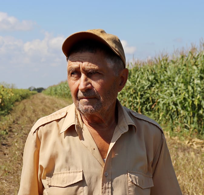 Elderly male farmer wearing a beige shirt and cap standing in a field with tall crops under a clear sky.