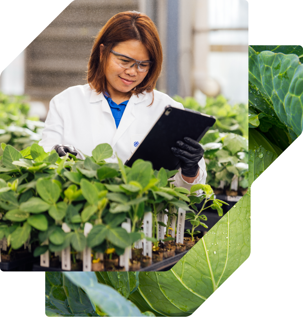 A female scientist in a white lab coat, safety glasses, and black gloves smiles while using a tablet to document data on leafy green seedlings in a greenhouse.