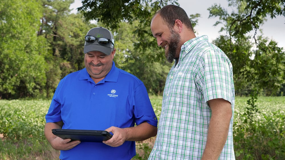Two men standing outdoors in a field, smiling while looking at a tablet device. One man is wearing a blue polo shirt and sunglasses, and the other is wearing a plaid shirt.