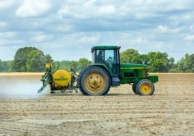Green tractor spraying a field with agricultural equipment labeled 'Redox' on a sunny day, with trees in the background.