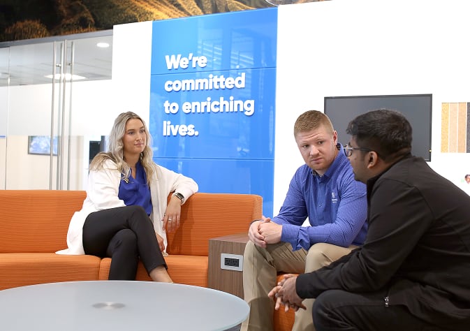 Three professionals having a discussion in a modern office space with a sign in the background that reads 'We're committed to enriching lives.