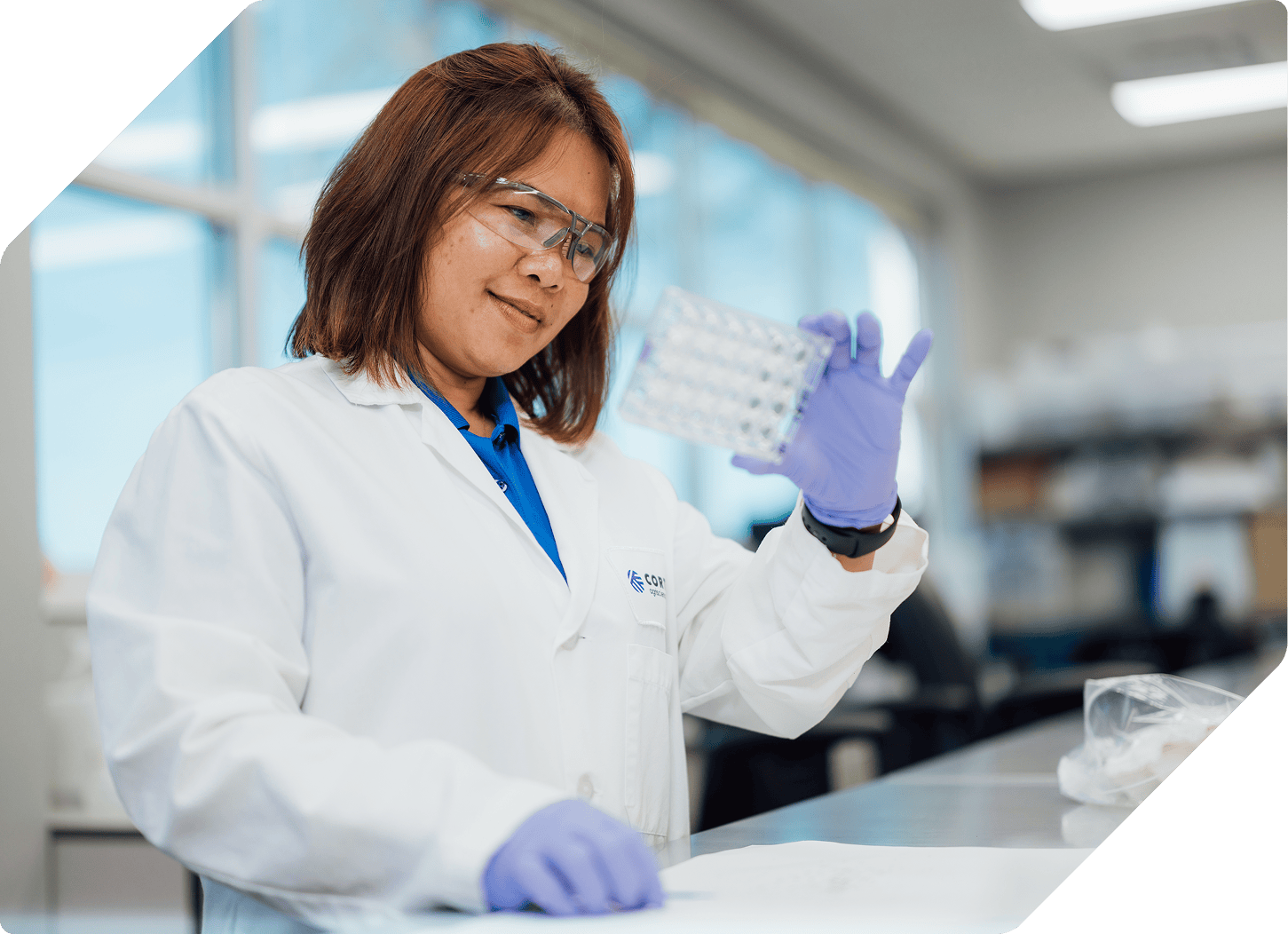 A female scientist in a white lab coat and safety goggles smiles while inspecting a multi-well laboratory plate in a bright, modern lab setting.