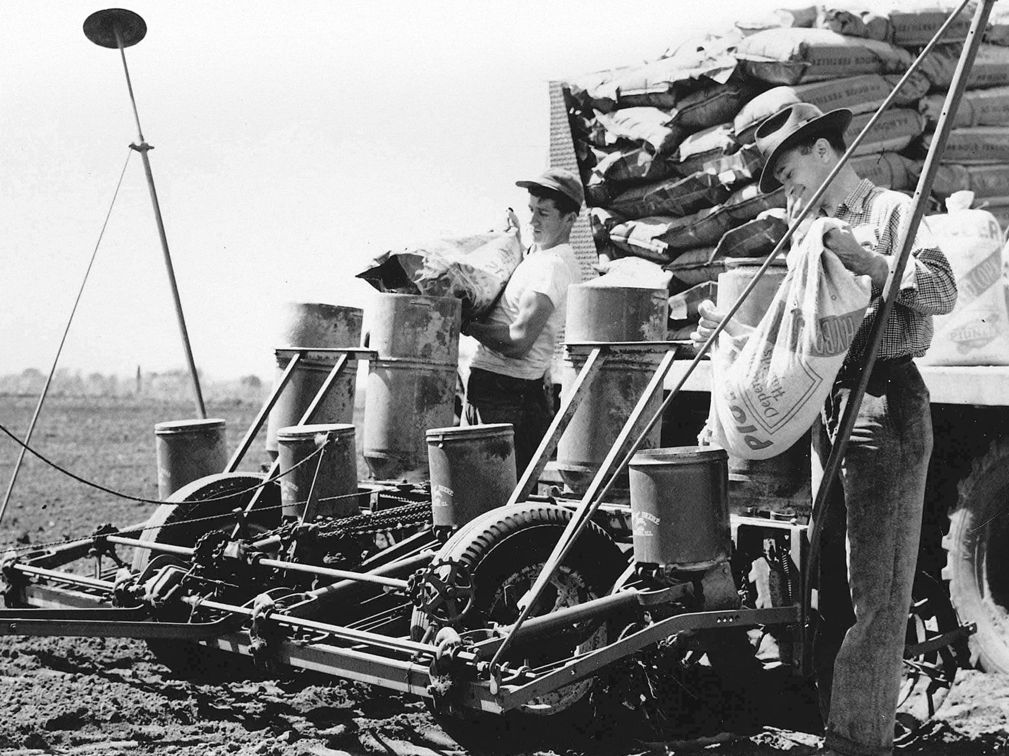 A black-and-white photo of two men loading large bags of seed into a vintage four-row trailing planter in a field.