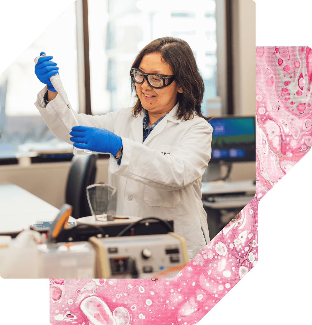 A smiling female scientist in a white lab coat, safety glasses, and blue gloves uses a pipette in a laboratory setting.