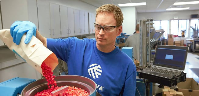 Laboratory technician wearing gloves and safety glasses pouring red coated seeds from a paper bag into a testing machine in a lab setting.
