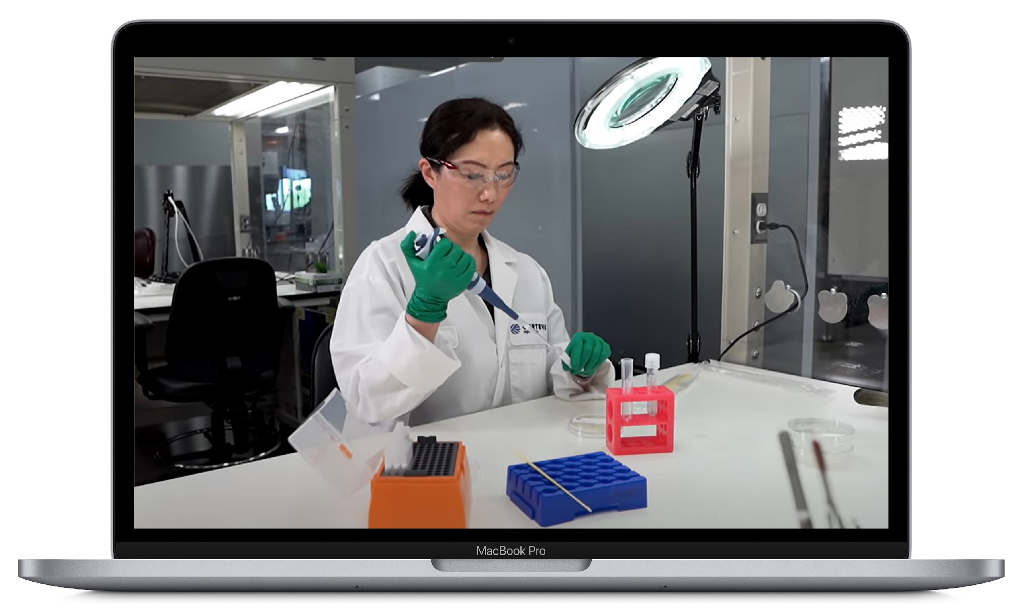 Scientist in a white lab coat and safety glasses using a pipette in a laboratory setting, shown on a MacBook Pro screen.