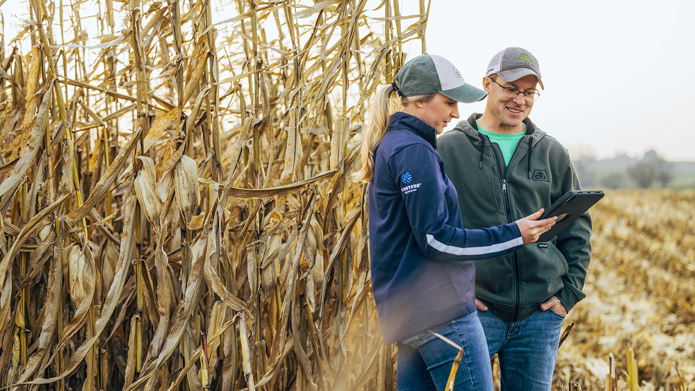 A woman in a navy jacket and a man in a green hoodie, both wearing baseball caps, stand in a field of dry corn while reviewing information on a tablet.