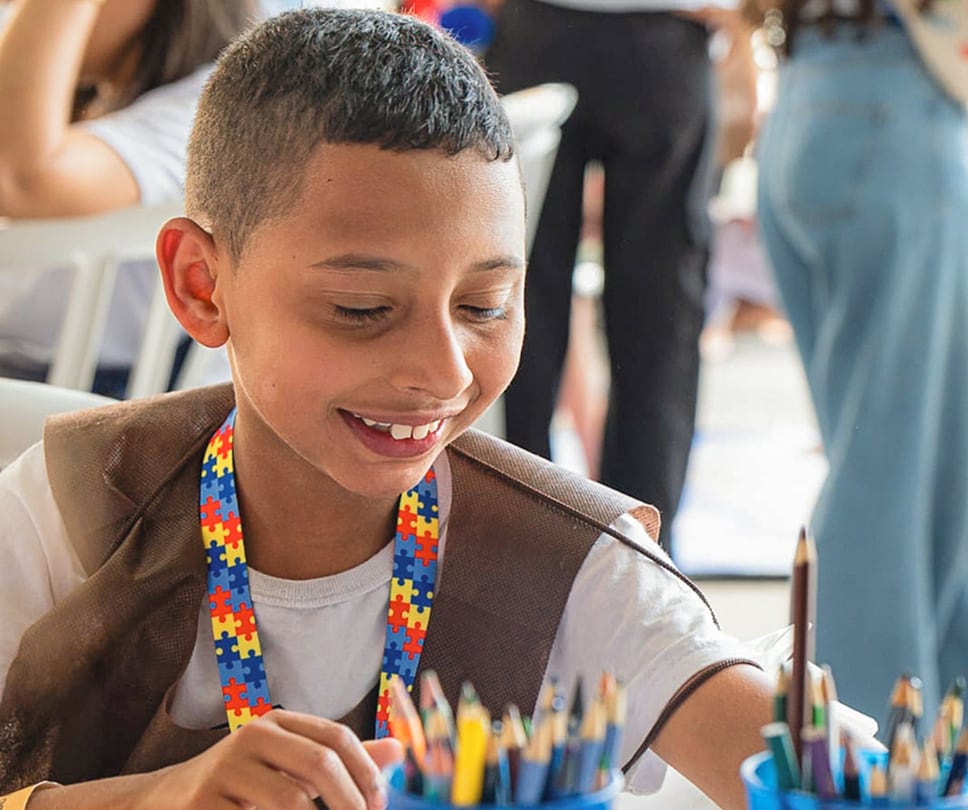 A young boy with a bright smile looks down at a collection of colored pencils. He is wearing a white shirt under a brown vest and a colorful lanyard.