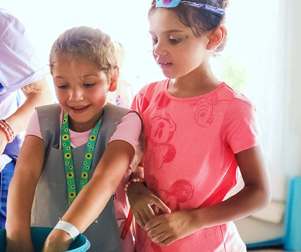 Two young girls look down with curiosity as one reaches into a blue bucket. One girl wears a sunflower-patterned lanyard.