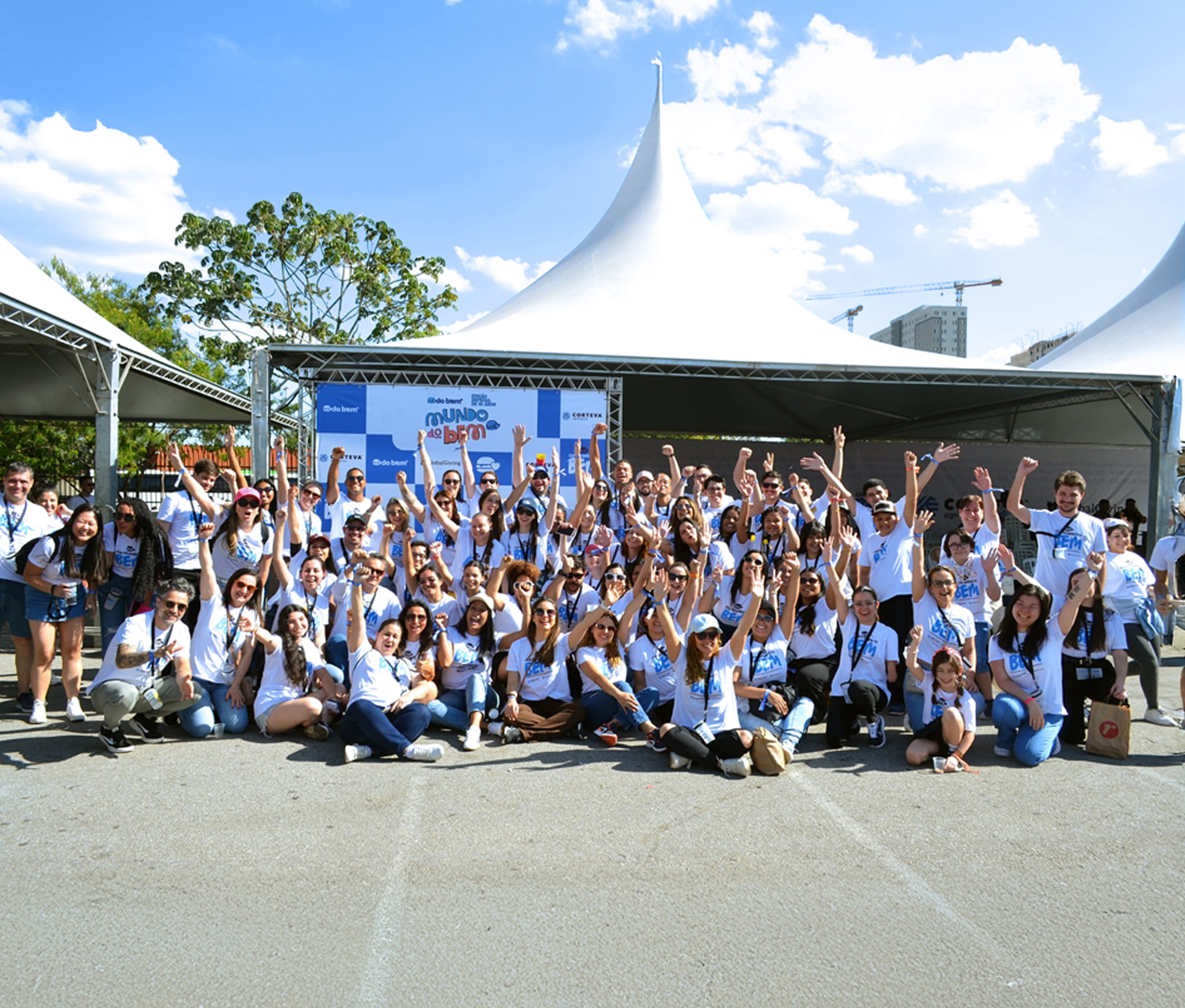 A large group of people in matching white "Mundo do Bem" t-shirts pose together outdoors, many with their arms raised in celebration.