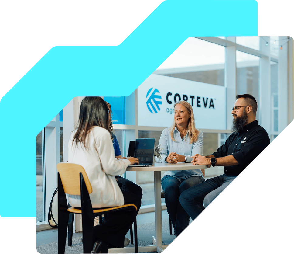 Four people sit around a small table in a modern office, engaged in a discussion. 2 laptops are open on the table, and the Corteva Agriscience logo is visible in the background.