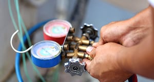 Close-up of a technician's hands adjusting a brass manifold gauge set with red and blue pressure dials, used for refrigeration servicing.