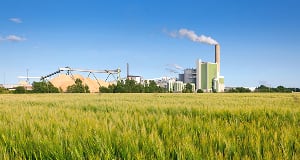 An industrial plant with a tall smokestack emitting white smoke, situated behind a lush green field under a clear blue sky.